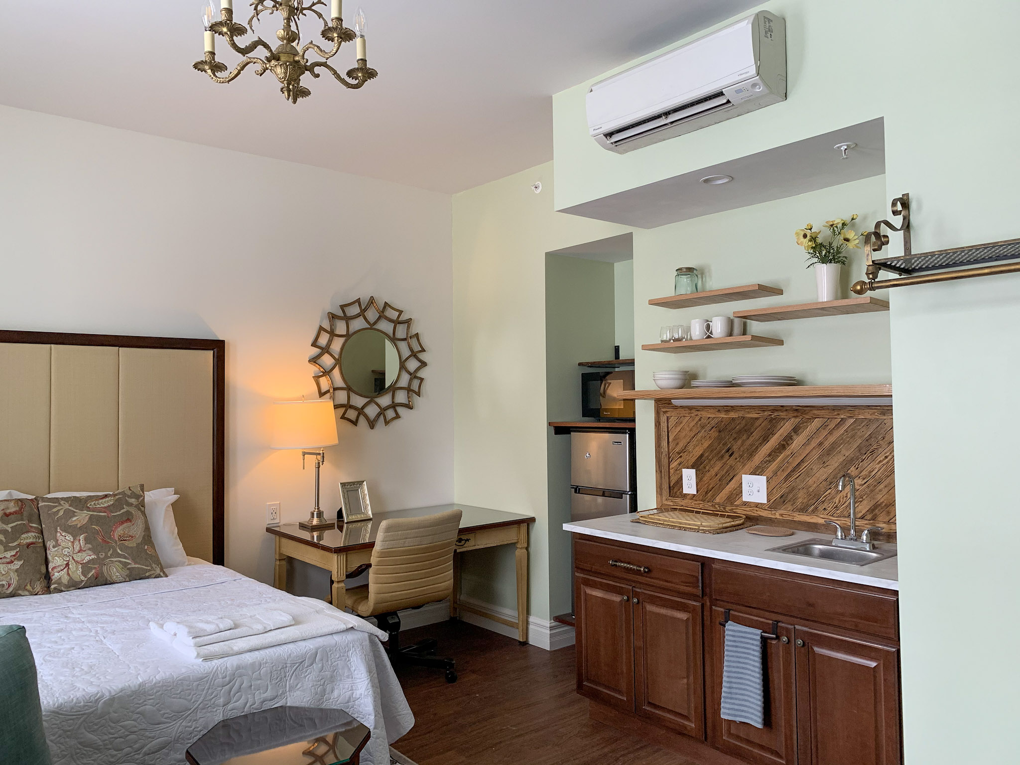 A furnished studio at Oaklands on 9th with a bed and folded towels, a desk and chair, and a kitchen area with wood cabinets and shelves.