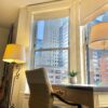 A wooden desk and cream office chair in a furnished studio's bay window at Oaklands on 9th, downtown Minneapolis towers outside.