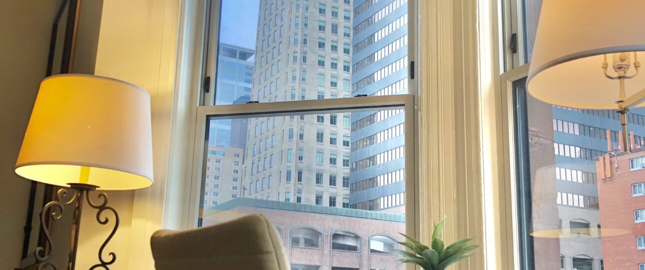 A wooden desk and cream office chair in a furnished studio's bay window at Oaklands on 9th, downtown Minneapolis towers outside.
