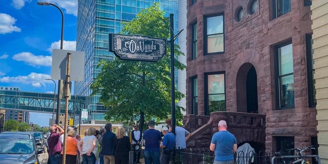 A crowd gathers on a sunny day outside the restored 1889 Oaklands on 9th brownstone in downtown Minneapolis for the historic designation plaque unveiling.