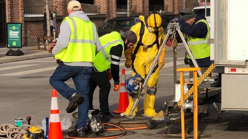 Minneapolis city crew in a yellow confined-space suit at a tripod winch over an open manhole in 9th Street, January 2021.