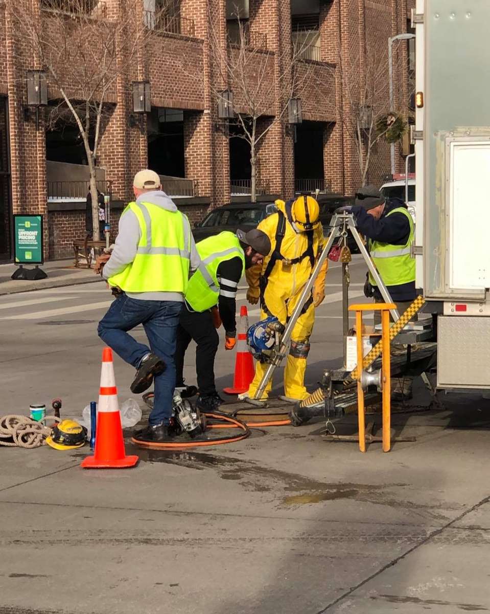 Minneapolis city crew in a yellow confined-space suit at a tripod winch over an open manhole in 9th Street, January 2021.